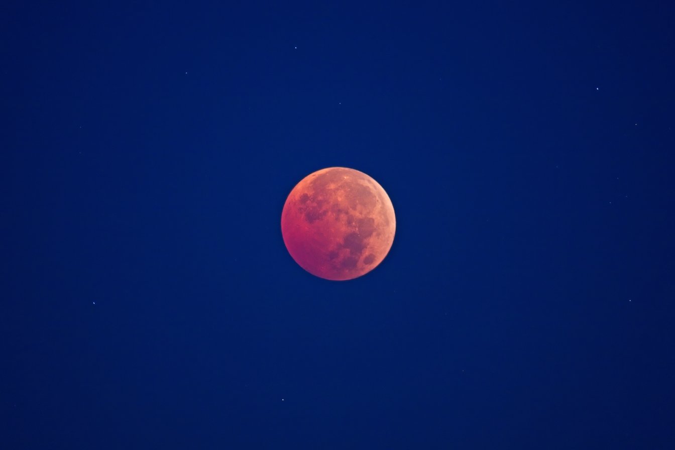 A total lunar eclipse, commonly known as a Blood Moon, featuring a large reddish-orange full moon set against a deep blue night sky. The moon's surface shows visible craters and maria, and a few faint stars are scattered across the dark sky.